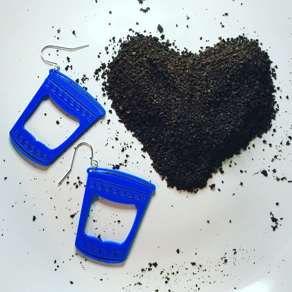 Two bright blue earrings that are shaped like iconic Greek to-go coffee cups in New York City. The NYC bodega cups are next to a heart formed from coffee grounds.