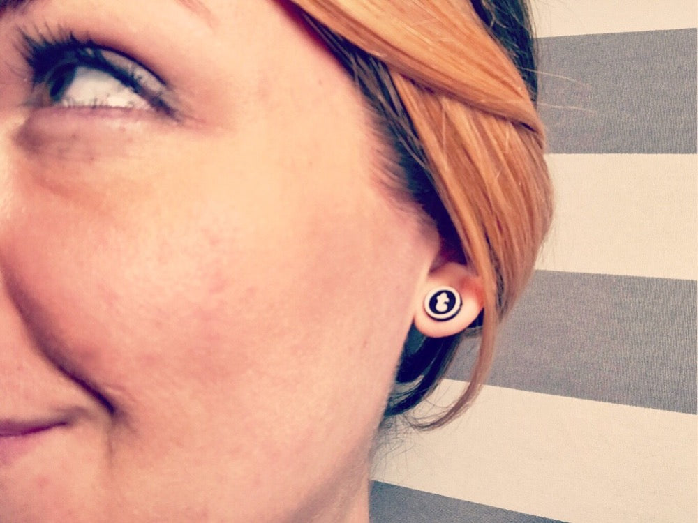 This is a close up of r+d's owner, Rebekah Thornhill wearing a 3D printed earring stud shaped like a typewriter key. It is a black circle with a white outline and a lowercase letter in the center. The letter shown here is t. 