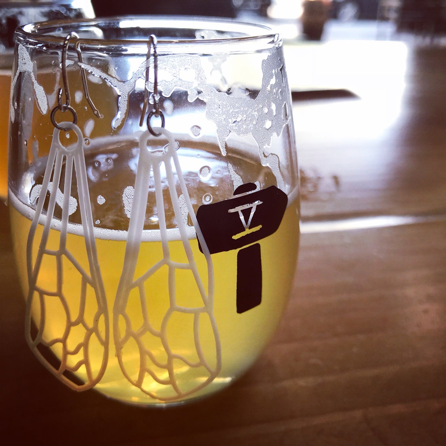 Hanging off of a small glass are two earrings that are shaped like dragonfly or insect wings. They are white and show all of the wing's veins. The glass has the logo of Fifth Hammer Brewery and is half full with a golden colored beer. 