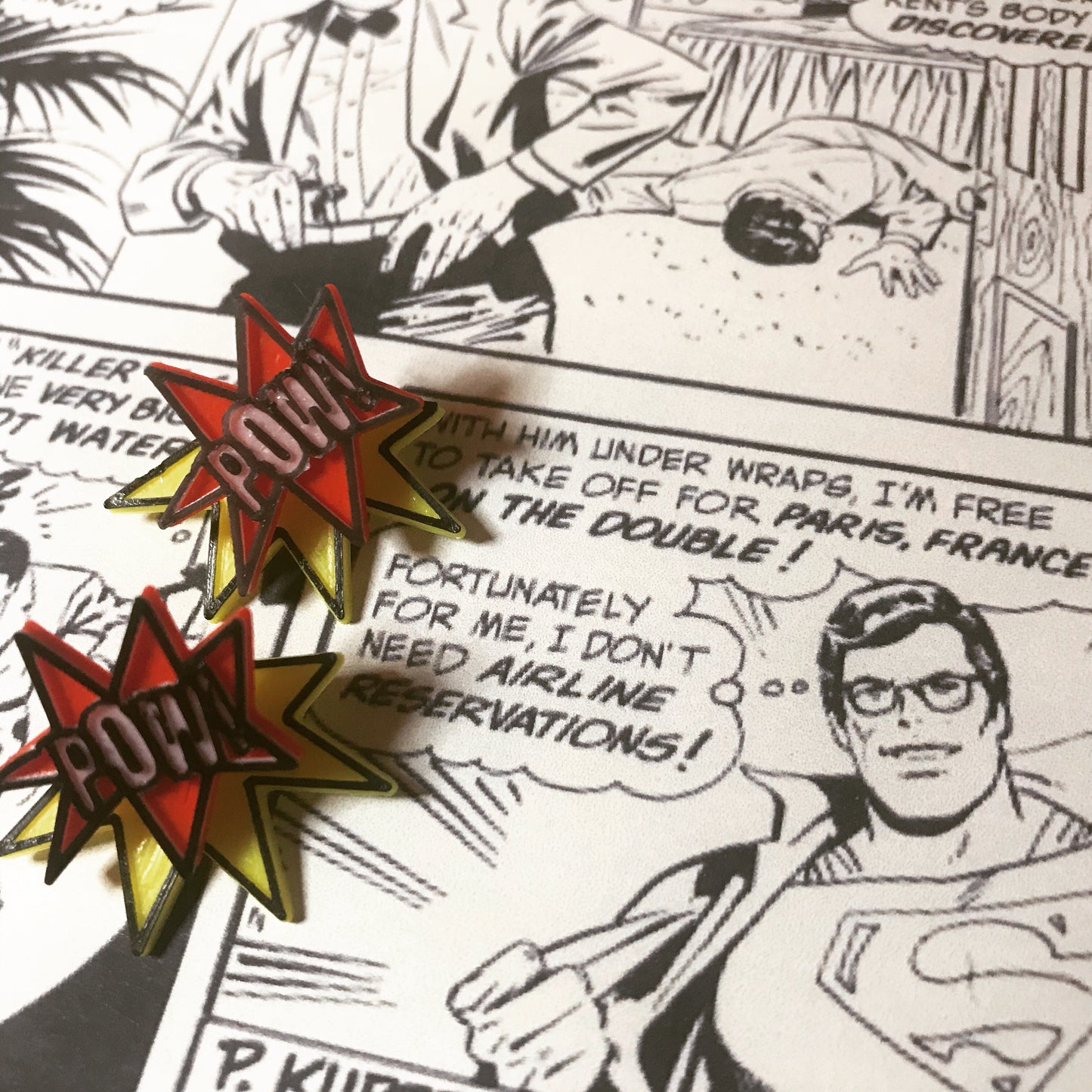 On a black and white comic book page with Superman/Clark Kent are two R+D earrings. They are front back earrings or ear jackets where there is a piece that hugs each side of the earlobe. The front shows a ref star burst that reads POW! in white. It is all outlined in black. The piece that fits behind the ear is a yellow starburst with a black outline. They are meant to look like a classic comic book illustration of a sound.