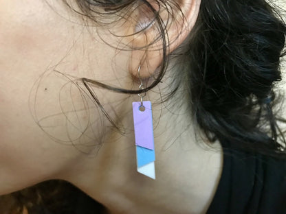 Show hanging from a woman's ear is an earring that is 3D printed with a plant based filament to make them sustainable as well as affordable. There are three banners hanging down; one is white, one is light blue, and the final is light purple. They are each different lengths so that the colors show.