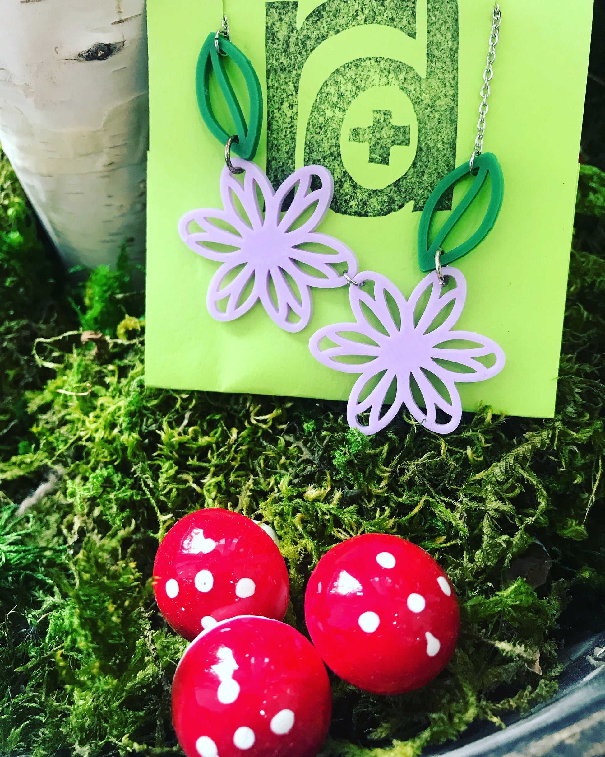 Sitting in a bed of green moss with red and white mushrooms is an R+D necklace on a card. The necklace is 3d printed and has two light purple flowers and two kelly green  leaves. 