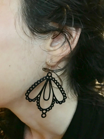 A close up of a woman with curly hair's ear and the black earring she is modeling. The earring  has multiple tear drop shapes that are accented with more circles and loops to look like lace.