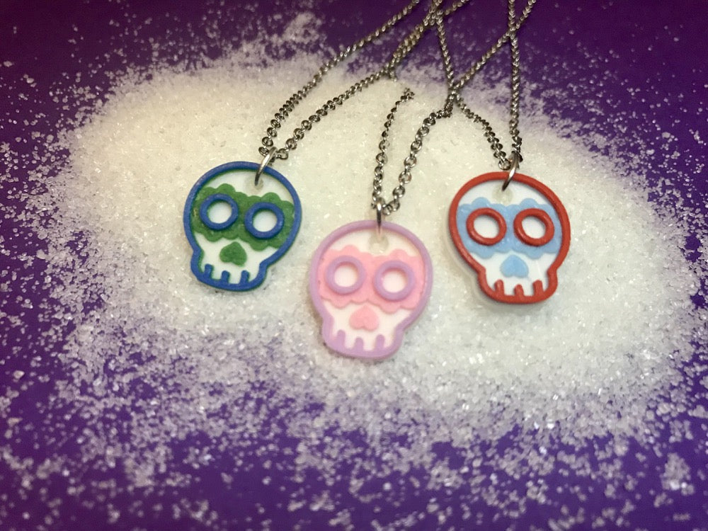 On a bright purple background there is a small pile of granulated sugar. Laying across the sugar are three necklaces; Each are 3D printed and in the shape of sugar skulls. One is white, green and blue; another is white, light pink and light purple; and the final is white, light blue, and red. The plant based filament used to print them is often made from sugar cane.