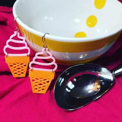 Two quirky earrings are hanging off of an empty bowl and next to a ice cream scoop. The earrings are made of a plant based polymer and have pink swirled soft serve on top of an orange sugar cone. 