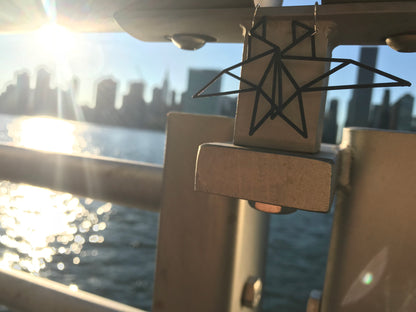 With the sun setting on the manhattan skyline in the background, there are two 3D printed R+D earrings hanging from the railing. They are both black and in the shape of geometric birds. 