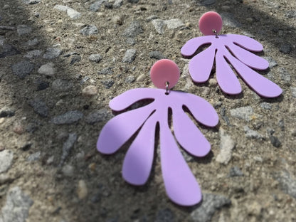 Resting on a sidewalk are two R+D earrings: they have a circlar piece at the top linked to an abstract shape based on Matisse's cut out work. This set is shown in light pink and light purple plant based filament. 
