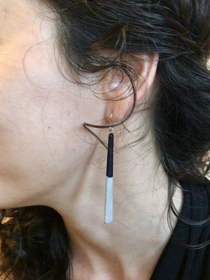 A close up shot of an ear and curly hair. Hanging from her earlobe is a  long minimalist dangle earring. They are a black plant based material that have been dipped into a metallic silver paint to make them shimmery.
