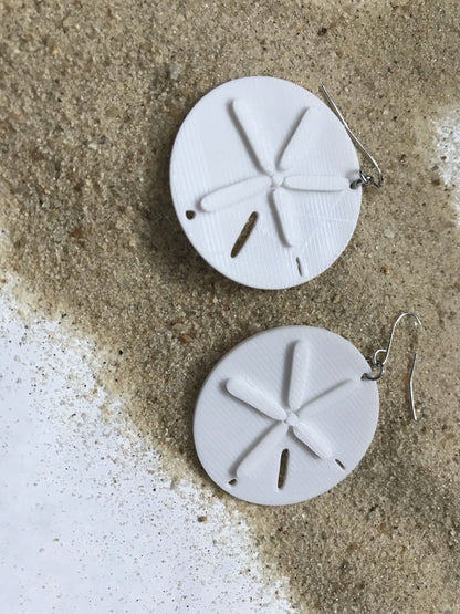 Tan sand is spread out with two earrings laying on top. The earrings are white and shaped like sand dollars.