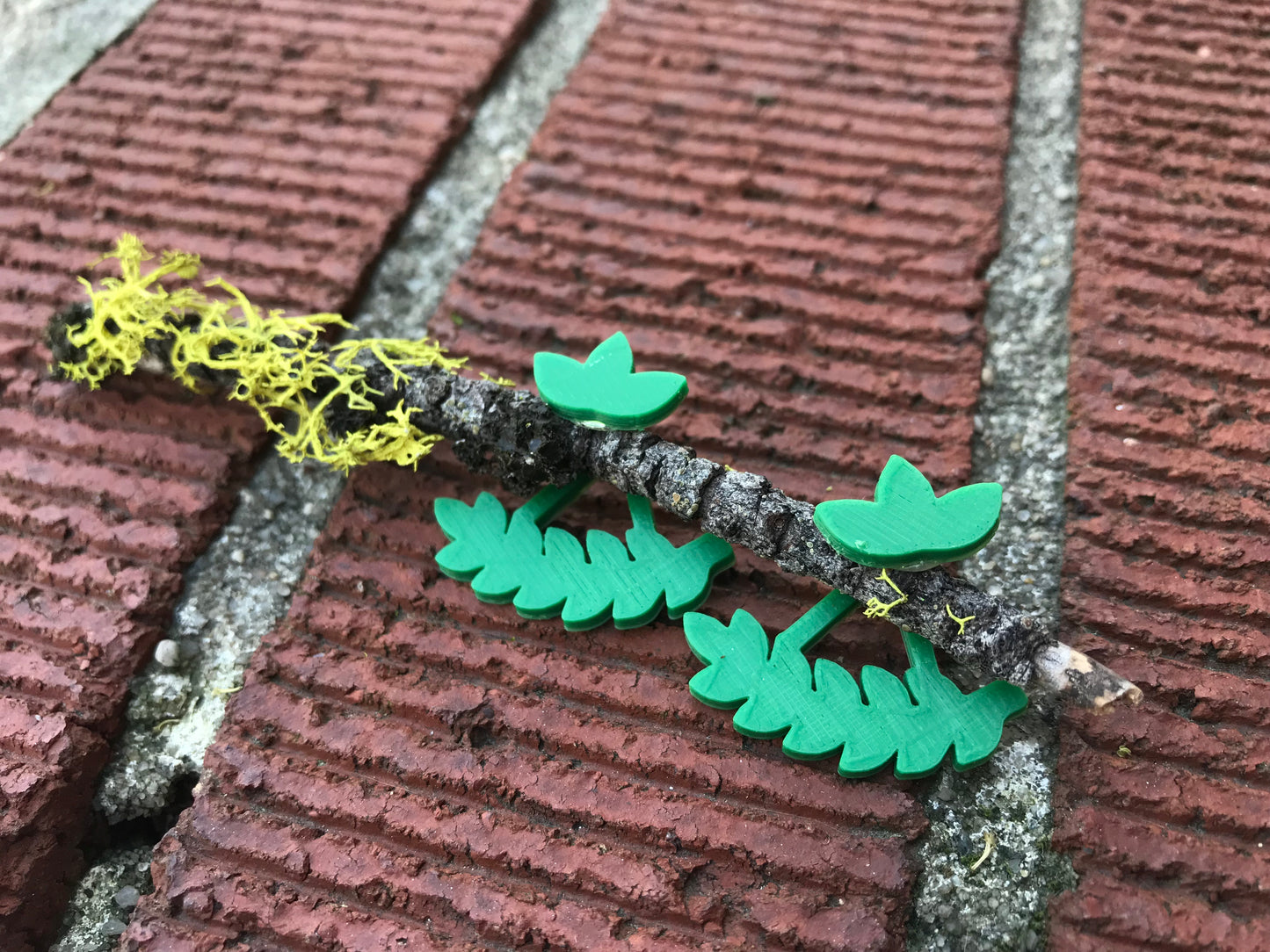 Two kelly green earrings are shown with a twig on a bricks. The earrings are front back earrings, so the twig is places between the front stud with a cluster of three leaves. The piece that fits behind the ear is behind the twig and shaped like a leafy bough or vine that curves with the shape of one's earlobe. These eco friendly 3D printed studs from R+D.