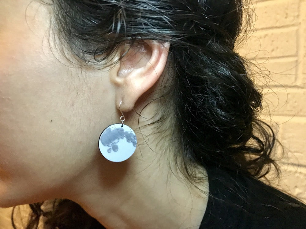 This is a close up shot of a woman wearing full moon earrings that have been 3D printed. They are bright white with silver accents to show the craters of the moon's surface. 