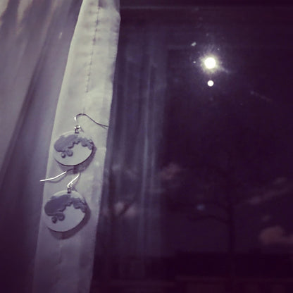 Hanging off of a white curtain are two plant based earrings. They have earring hooks with two circle moons hanging beneath. The moons are white with silver accents to show the craters of the moon's surface. In the background is a window with the night sky and a bright moon shining back. 