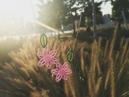 With a sunset, wheat, and trees in the background, an R+D necklace is held in the frame. The necklace has light purple flowers and kelly green leaves. 