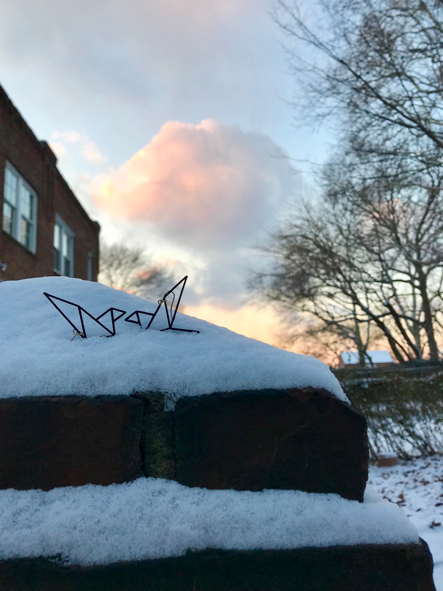 There is a fresh snowfall with barren trees and brick houses in the background. The sky is a bright blue with a cloud reflecting pinks and yellows of a setting sun. In a pile of snow are two geometric bird earrings from R+D they are black and peaking out of the pile as though they had just landed on that spot.
