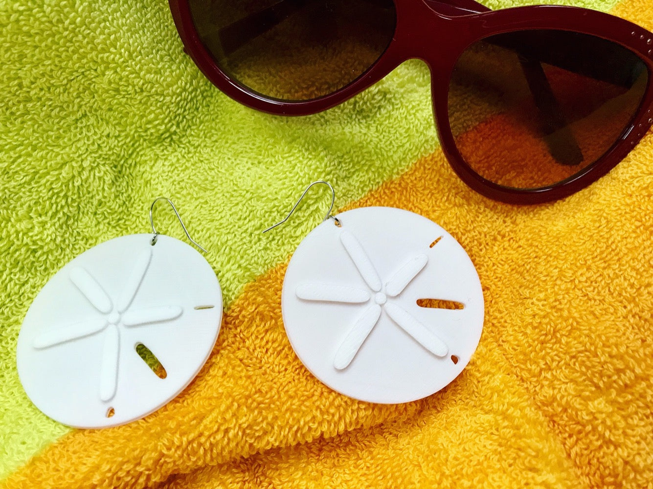 Laying on a beach towel and next to sunglasses are two white earrings shaped like sand dollars or silver dollars that have washed onto shore. 