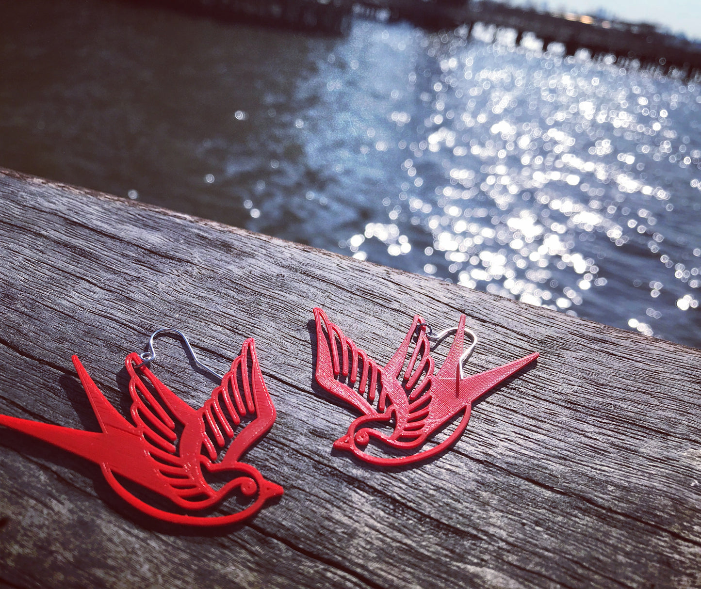 Shown on a wooden pier with the shimmering water in the background are two 3D printed R+D earrings. They are a bright fire engine red color and shaped like  swallows or sparrows. The birds are a classic look like sailor tattoos that mark one's journey. 