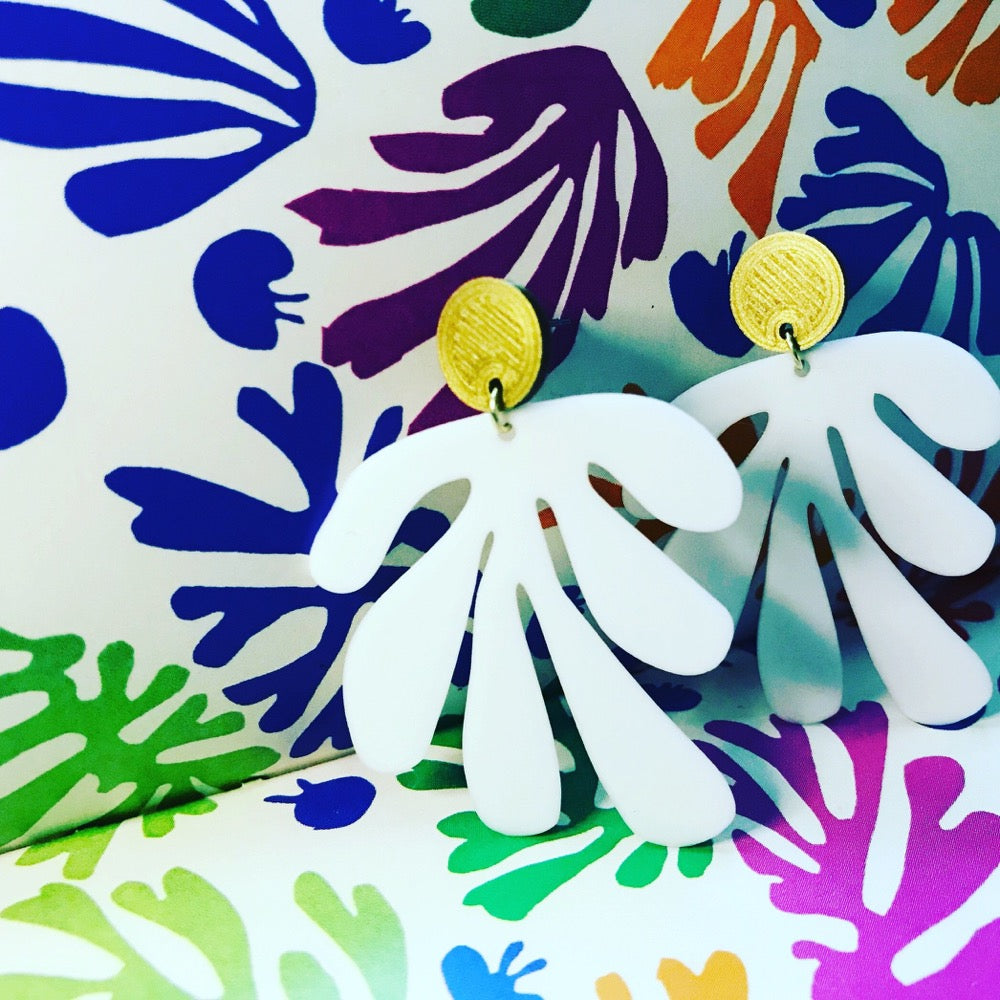 Shown in a box with Matisse's cut out shapes, there are two earrings standing up. The earrings have a circlar piece at the top linked to an abstract shape based on Matisse's cut out work. This set is shown in gold and white plant based filament. 
