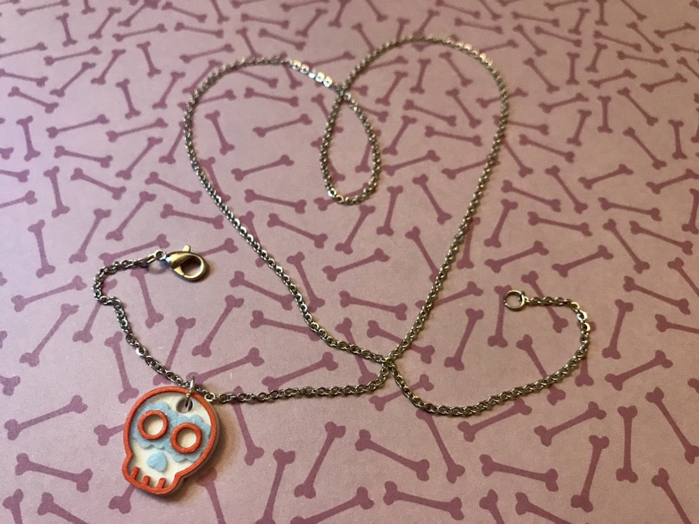 On a purple background with a pattern of bones is a necklace. The chain is laid and twisted into a heart shape with a 3D printed pendant in the shape of a sugar skull. 