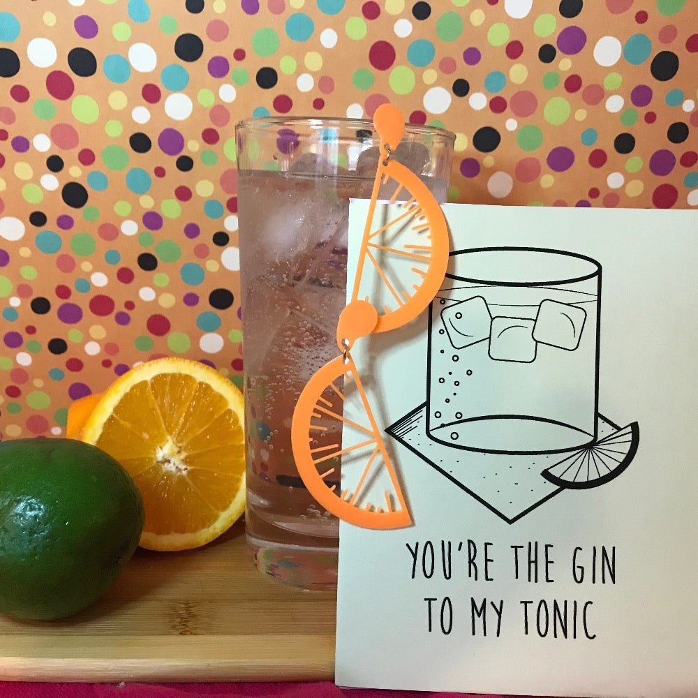 In front of an orange background with multicolor dots .is a cutting board with citrus, a tall glass with ice, gin, and tonic. Hanging off the glass are two R+D earrings. They are orange and shaped like citrus slices with drips of juice. There is also an R+D card that has a glass, ice, napkin and fruit illustrated with the line, You're the Gin to my tonic. 