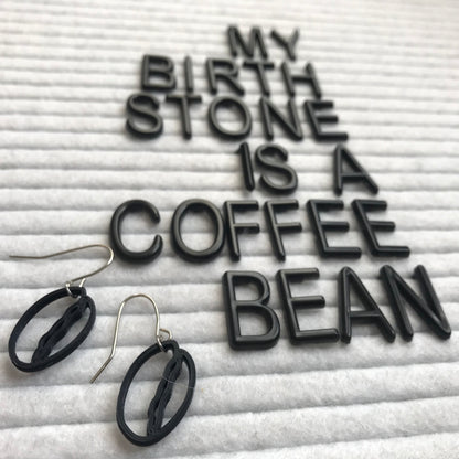 There is a felt letter board that reads, my birthstone is a coffee bean and in the foreground two black plant based earrings that are shaped like coffee beans. 
