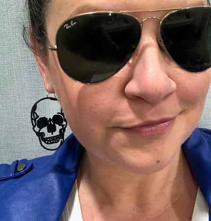 A woman wearing Ray Ban aviator sunglasses and a blue leather jacket smirks at the camera. You can see her skull earring, which forms a perfect black silhouette as it dangles from her earlobe. 