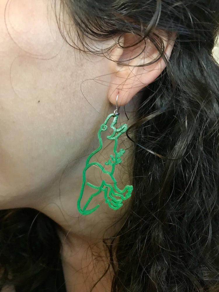 This is a close up of a woman's ear. She is wearing a 3D printed designed to look like a dinosaur. The T-Rex shape hangs from the hook as though the dinosaur were reaching to bite the earlobe. It is made with our bright kelly green plant based filament. 