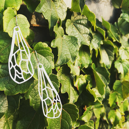 In the background is a wall covered in lush green vines. In the forefront are two white wing earrings. The earrings look like dragonfly or insect wings that have all of the veins visible. 
