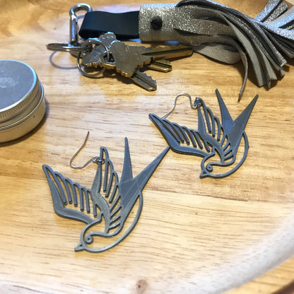 On a wooden catch all dish are a set of keys with a silver leather tassel keychain, an aluminum tin and two silver R+D 3D printed earrings. The earrings are birds that look like they are swooping down. They are reminiscent to classic sailor tattoos of swallows. 