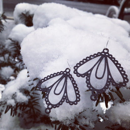 Two earrings are shown on a crisp white snow that has freshly gathered on a bush. The earrings are both black and look like lace with three teardrop shapes.