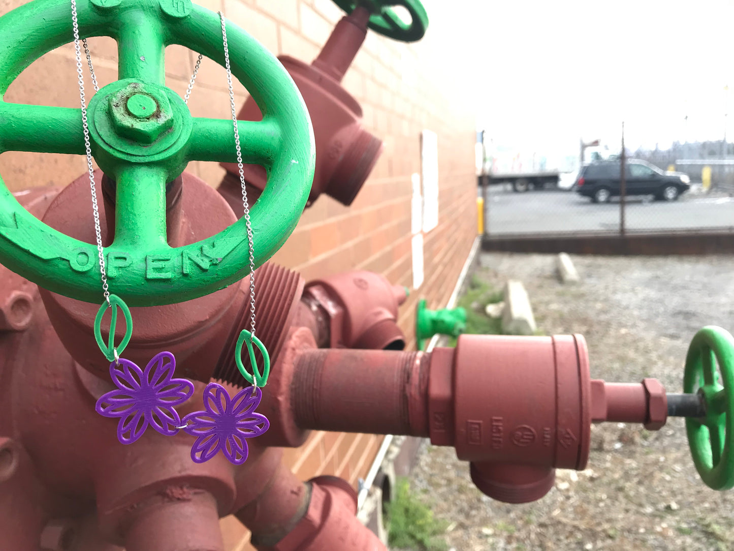 Hanging off of a cluster of rust red pipes and green handles is a 3D Printed R+D necklace. It is asymmetrical with two bright purple flowers and two kelly green leaves. 