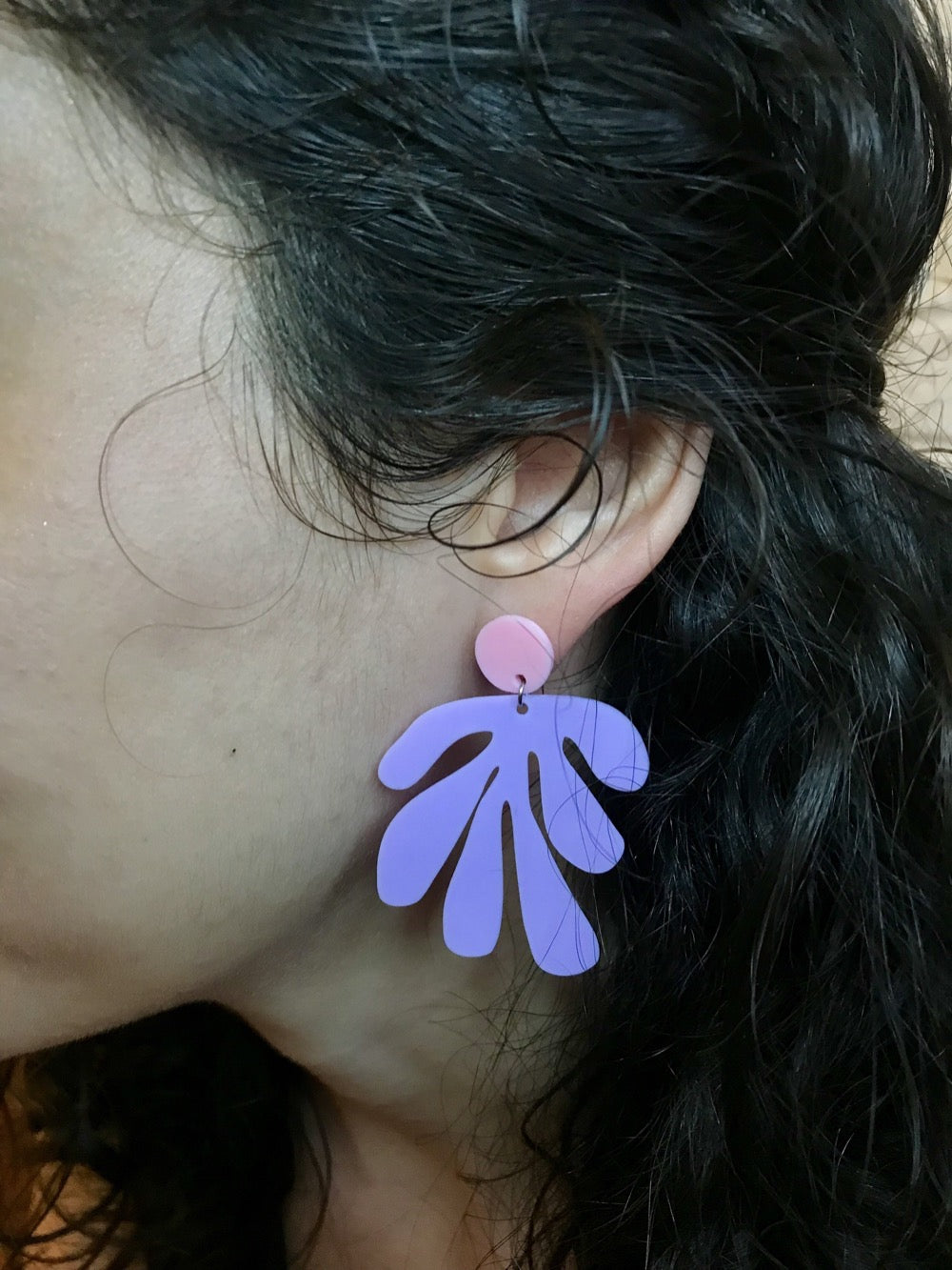 This is a close up of a woman's ear. She is wearing an R+D earring that has a circlar piece at the top linked to an abstract shape based on Matisse's cut out work. This earring is shown in light pink and light purple plant based filament. 