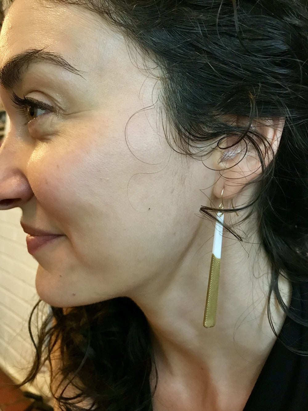 A woman with curly hair smiles as she models long minimalist dangle earrings. They are a white plant based material that have been dipped into a metallic gold paint to make them shimmery. 
