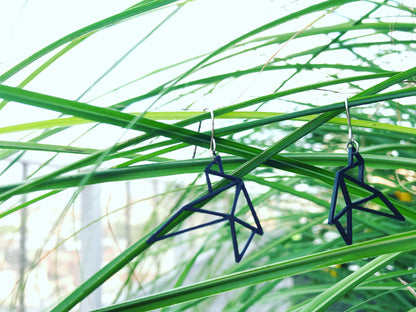 Two R+D earrings are shown hanging off of saw grass with a bright background that is blurred. The earrings are 3d printed in an eco friendly black filament and are shaped like two delicate geometric birds in flight.