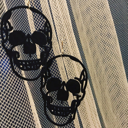 Two earrings are shown on thin, light curtains. The black earrings are shaped like human skulls and are realistic, edgy, and look like they could be smiling. 