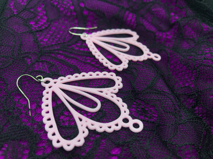 Laying on magenta and black lace are two earrings they are light pink and have large teardrop shapes that come together to make a lacy form. 