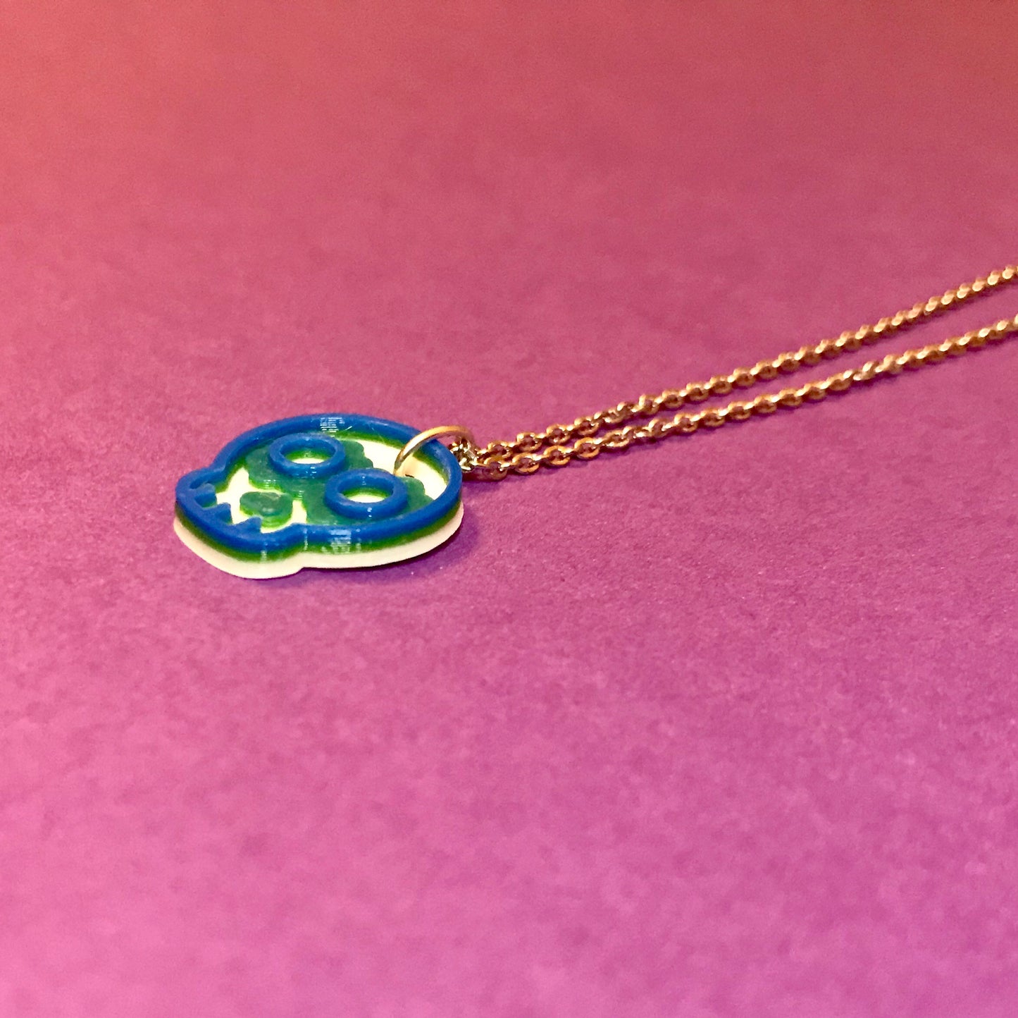 In the center of a purple background is a necklace laying down. It is 3D printed and laying so that you can see the layers of this design; first it is printed in white, next in green, and finished in blue. 
