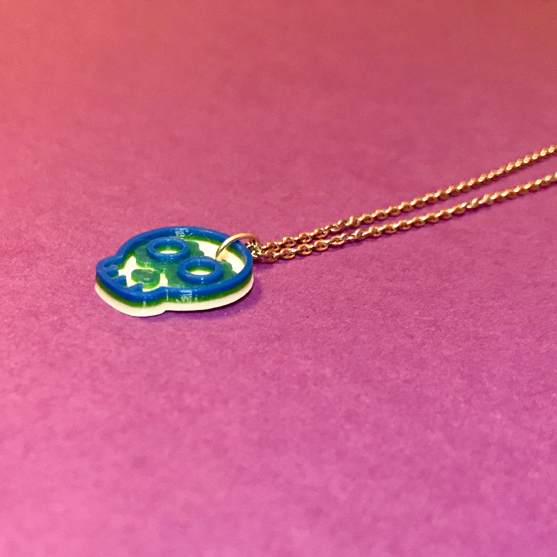 In the center of a purple background is a necklace laying down. It is 3D printed and laying so that you can see the layers of this design; first it is printed in white, next in green, and finished in blue. 