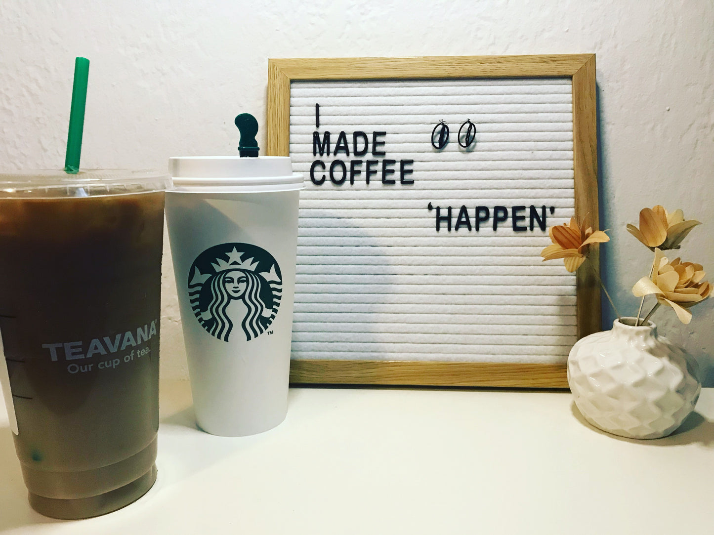 A letter board has the words, I made coffee 'happen'. Also hanging on the letter board are two black earrings shaped as coffee beans. In the foreground are two large Starbuck coffees and a small vase with wooden flowers. 