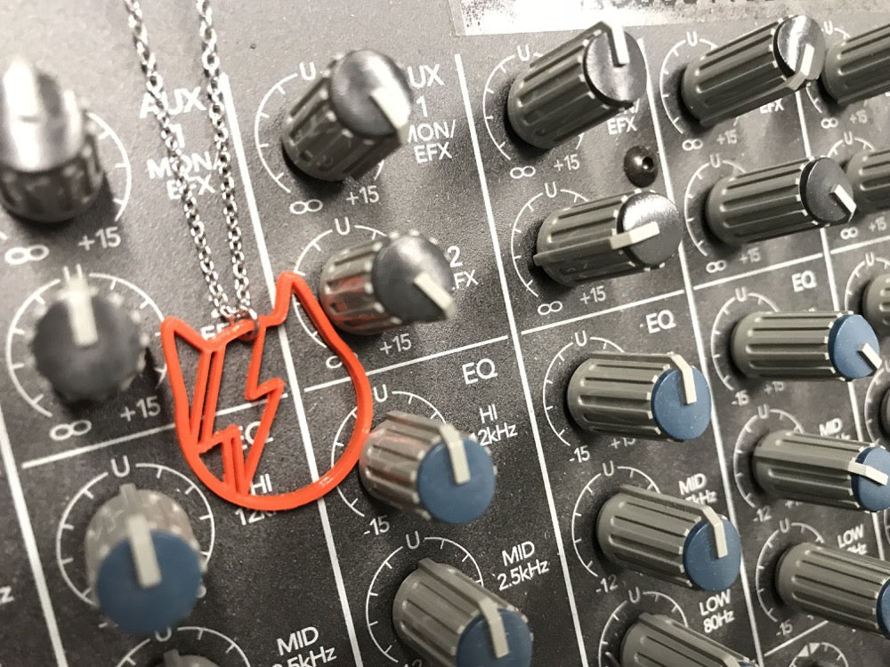 Hanging off of a sound board with lots of dials is a 3D printed R+D necklace. The necklace pendant is in the shape of a cat with pointing ears and has a lightning bolt across it's face to mimic the iconic David Bowie album cover. 