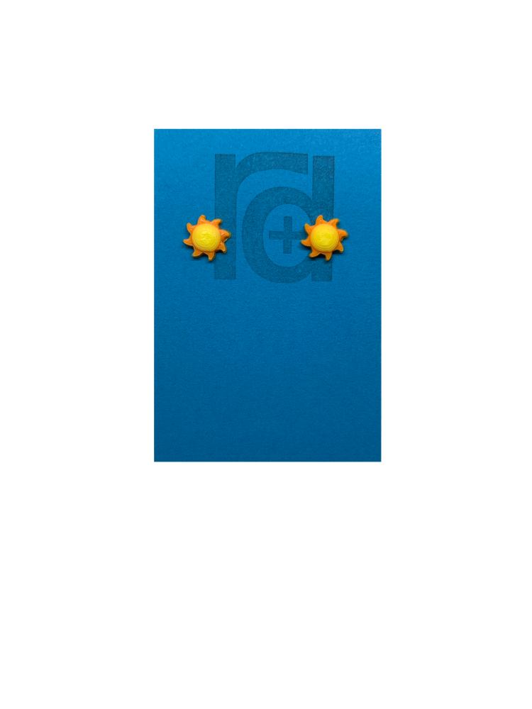 On a bright sky blue earring card with the R+D logo are two 3D Printed studs that are shaped like suns. They are very small, with round yellow centers and eight orange beams radiating out from the center. These are printed with an eco friendly material and are biodegradable.