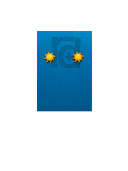 On a bright sky blue earring card with the R+D logo are two 3D Printed studs that are shaped like suns. They are very small, with round yellow centers and eight orange beams radiating out from the center. These are printed with an eco friendly material and are biodegradable.