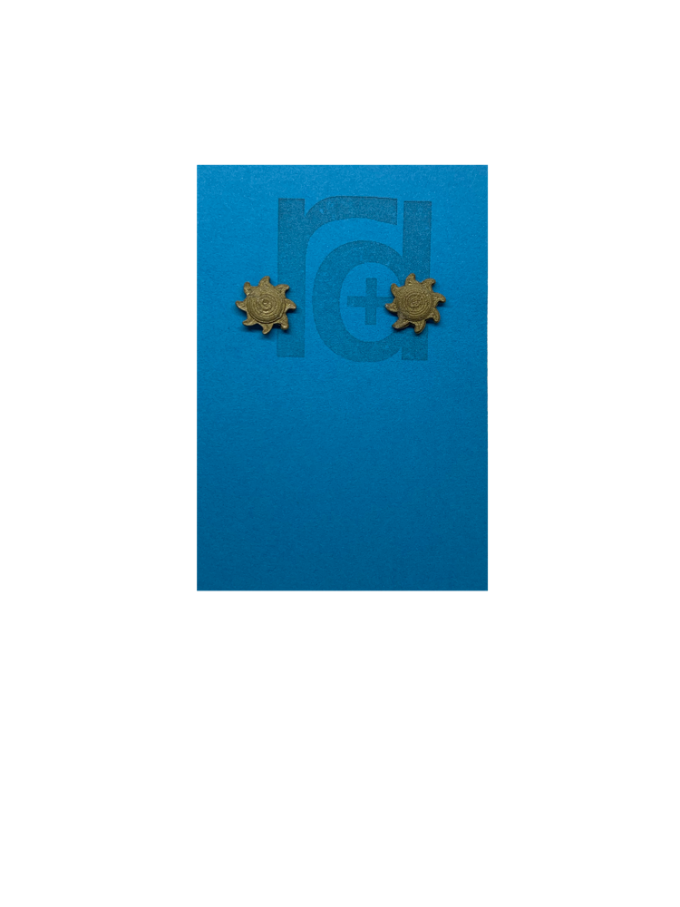 On a bright sky blue earring card with the R+D logo are two 3D Printed studs that are shaped like suns. They are very small and printed in a solid gold color. They have rounded centers and eight beams radiating out from the center. These are printed with an eco friendly material and are biodegradable.