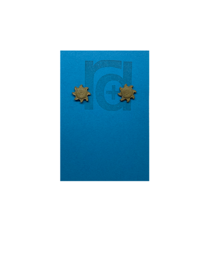 On a bright sky blue earring card with the R+D logo are two 3D Printed studs that are shaped like suns. They are very small and printed in a solid gold color. They have rounded centers and eight beams radiating out from the center. These are printed with an eco friendly material and are biodegradable.