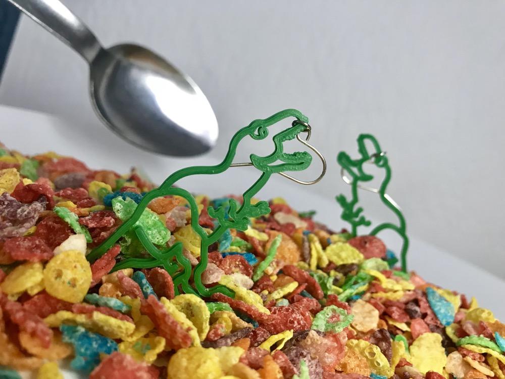In the foreground is a bowl of fruity pebbles cereal with a dinosaur earring istanding up in the cereal. There is a second earring in the background. Both dinosaur earrings are shaped like T-rexes and are a bright kelly green color. 