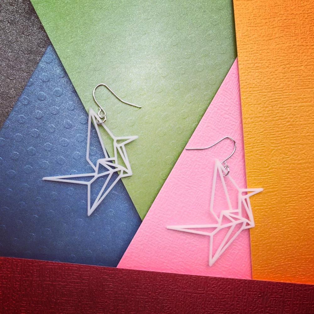 On a background of multiple brightly colored papers are two white 3D printed earrings. The earrings form the shape of folded origami cranes. 