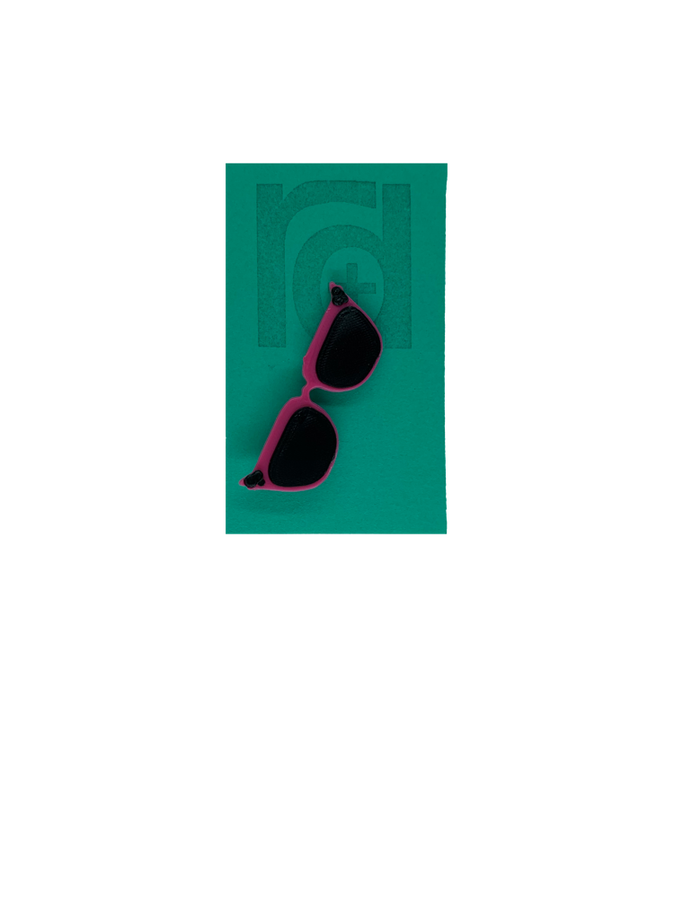 On a teal card is a R+D 3D printed pin. They're in the shape of cat eye sunglasses and have hot pink frames, black lenses, and black accents at the top corners of the glasses. 