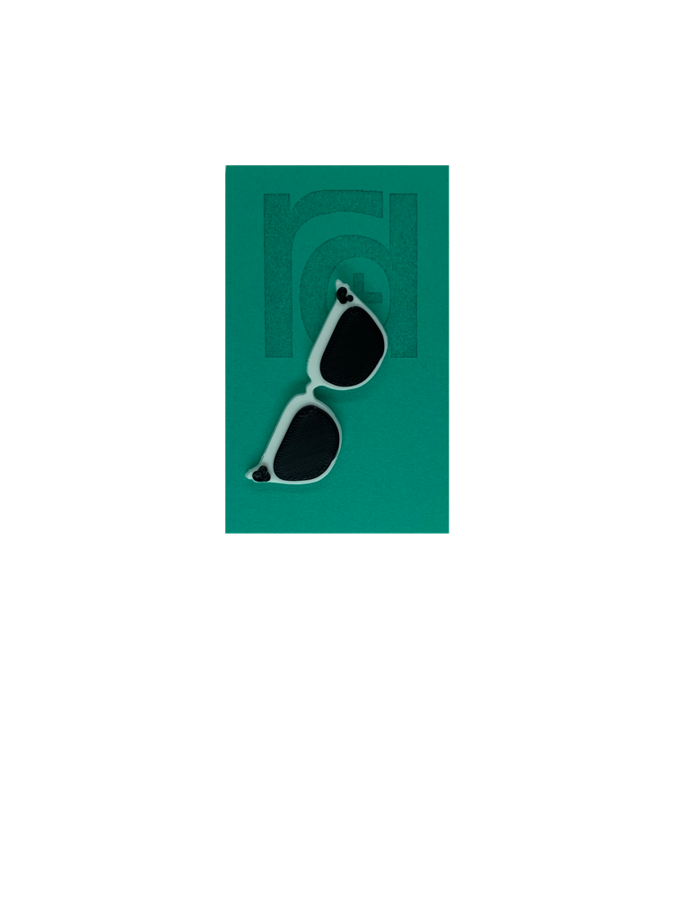 On a teal card is a R+D 3D printed pin. They're in the shape of cat eye sunglasses and have white frames, black lenses, and black accents at the top corners of the glasses.