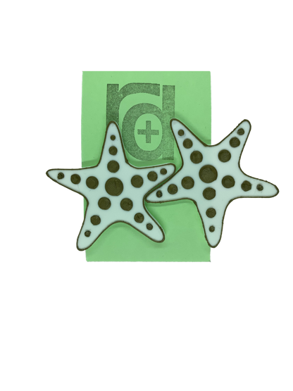 Hanging on a green earring card are two large R+D earrings shaped like star fish.  They are white with a gold outline and have circle accents across each arm and in the center.