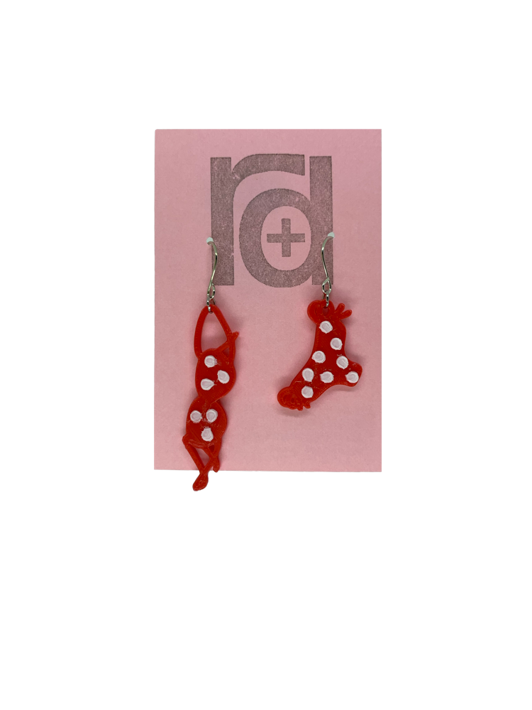 Hanging off of a pink earring card are two R+D earrings. They are asymmetrical  earrings shaped like a classic bikini  that is hung out to dry. The bottoms have ties on the sides. They are red with white polka dots.