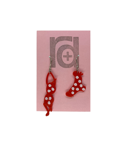 Hanging off of a pink earring card are two R+D earrings. They are asymmetrical  earrings shaped like a classic bikini  that is hung out to dry. The bottoms have ties on the sides. They are red with white polka dots.